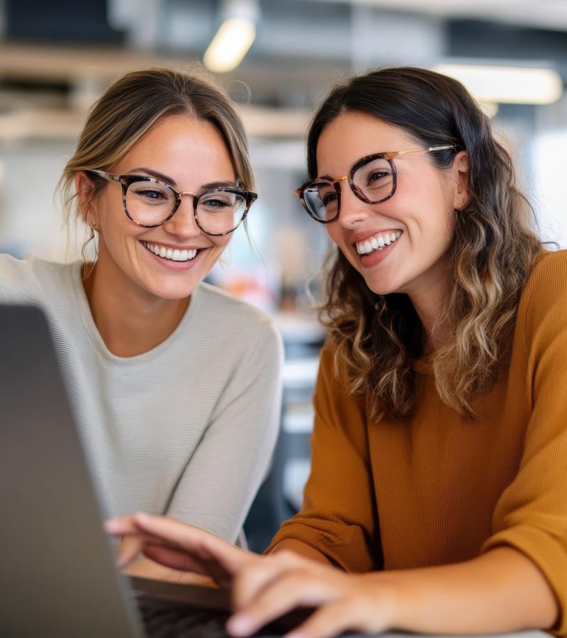 Two women wearing glasses and casual outfits are engaging in a collaborative project on a laptop in a bustling modern office, exuding teamwork and positivity.